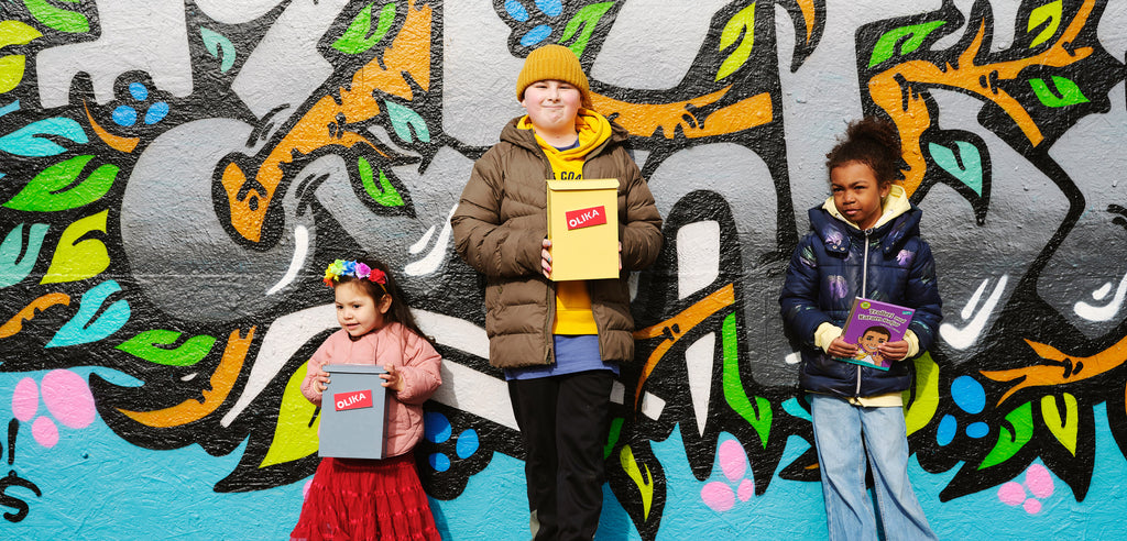 Tre barn står med varsin brevlåda framför en vägg med graffitimålning. 