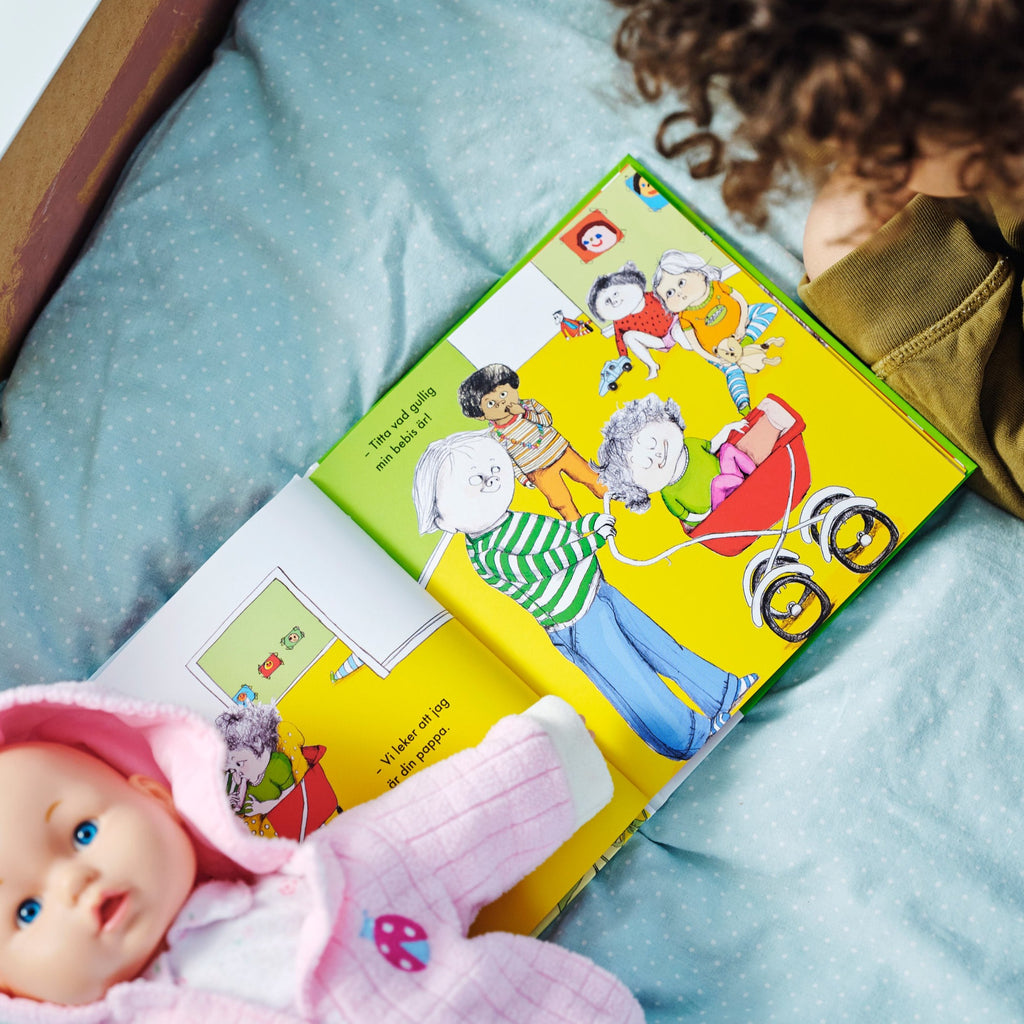 Ett barn ligger på en säng tillsammans med en rosaklädd docka och ett färgglatt uppslag i Vi leker att jag är din pappa - Bilderbok 1-3 år - OLIKA förlag - Författare: Tora von Platen - Illustratör: Emili Kvarnström Svensson