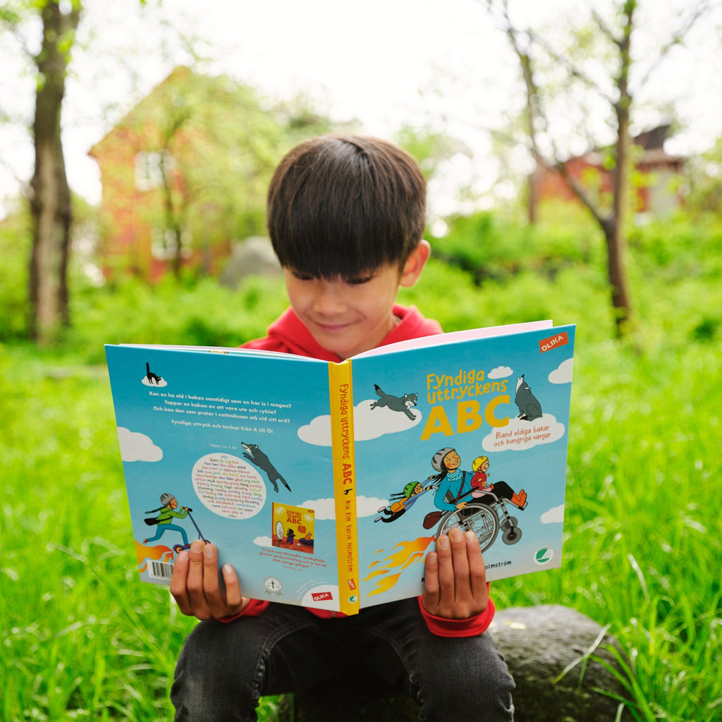 Ett barn sitter i gräset och läser Fyndiga uttryckens ABC - Bilderbok 3-6 år - OLIKA - Författare: Mia Kim - Illustratör: Karin Holmström