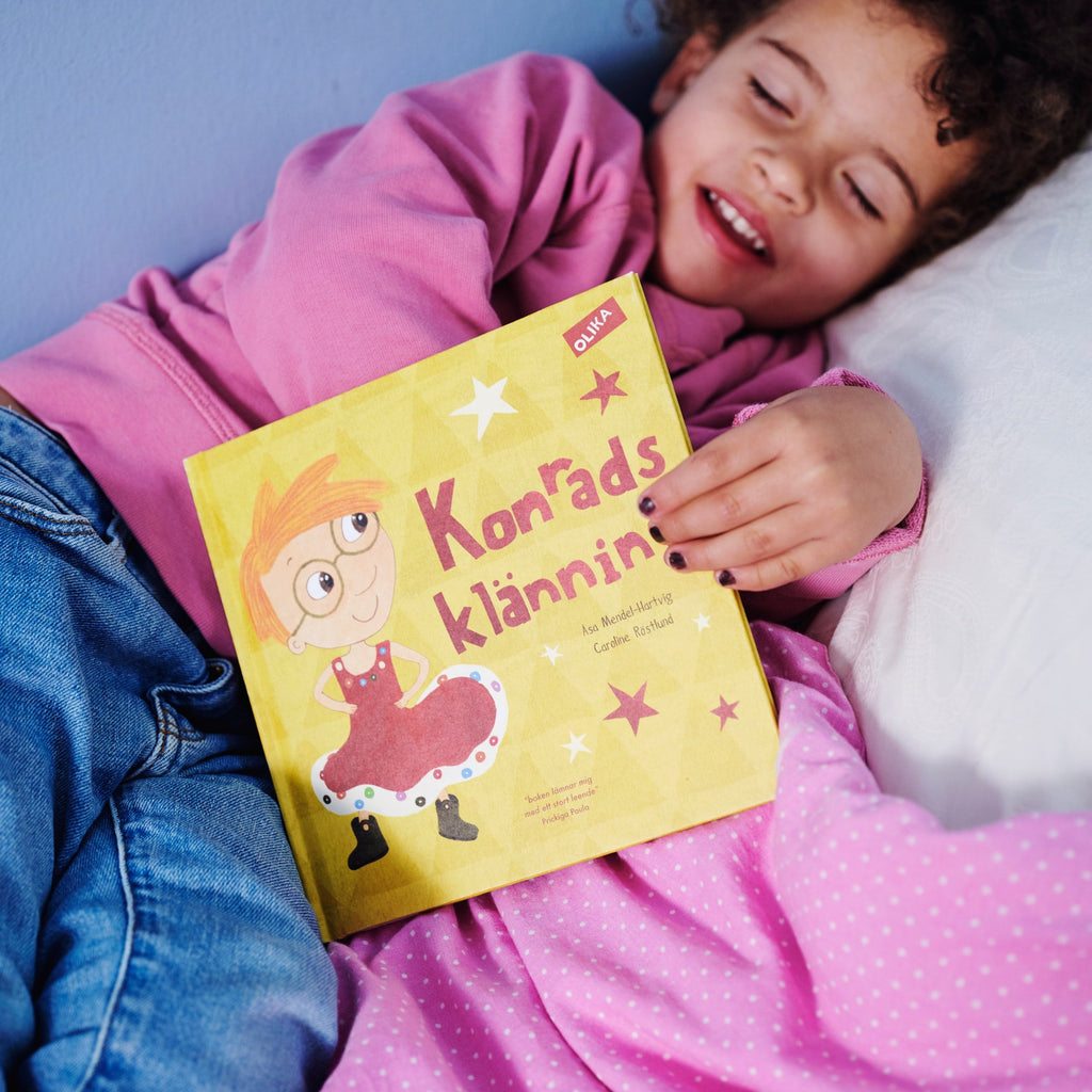 Ett barn i rosa tröja blundar och håller fram en kopia av Konrads klänning - Bilderbok 1-3 år - OLIKA förlag - Författare: Åsa Mendel-Hartvig - Illustratör: Caroline Röstlund