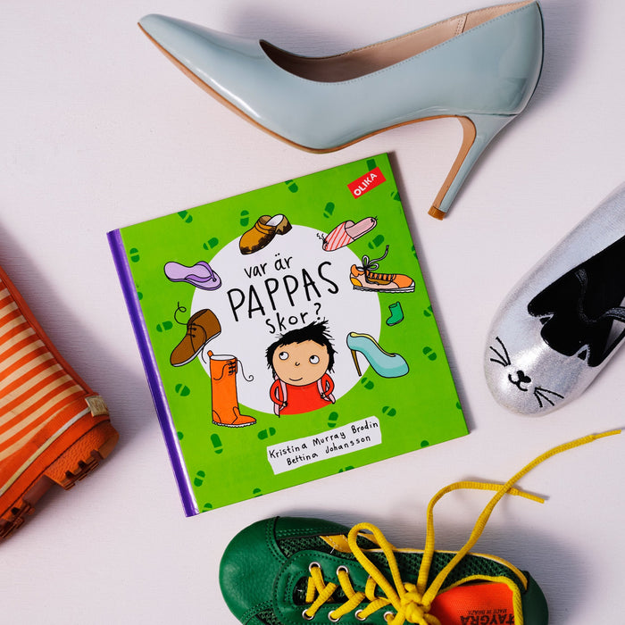 Bilderboken Var är pappas skor? syns på vit yta, omgiven av olika typer av skor i olika storlekar