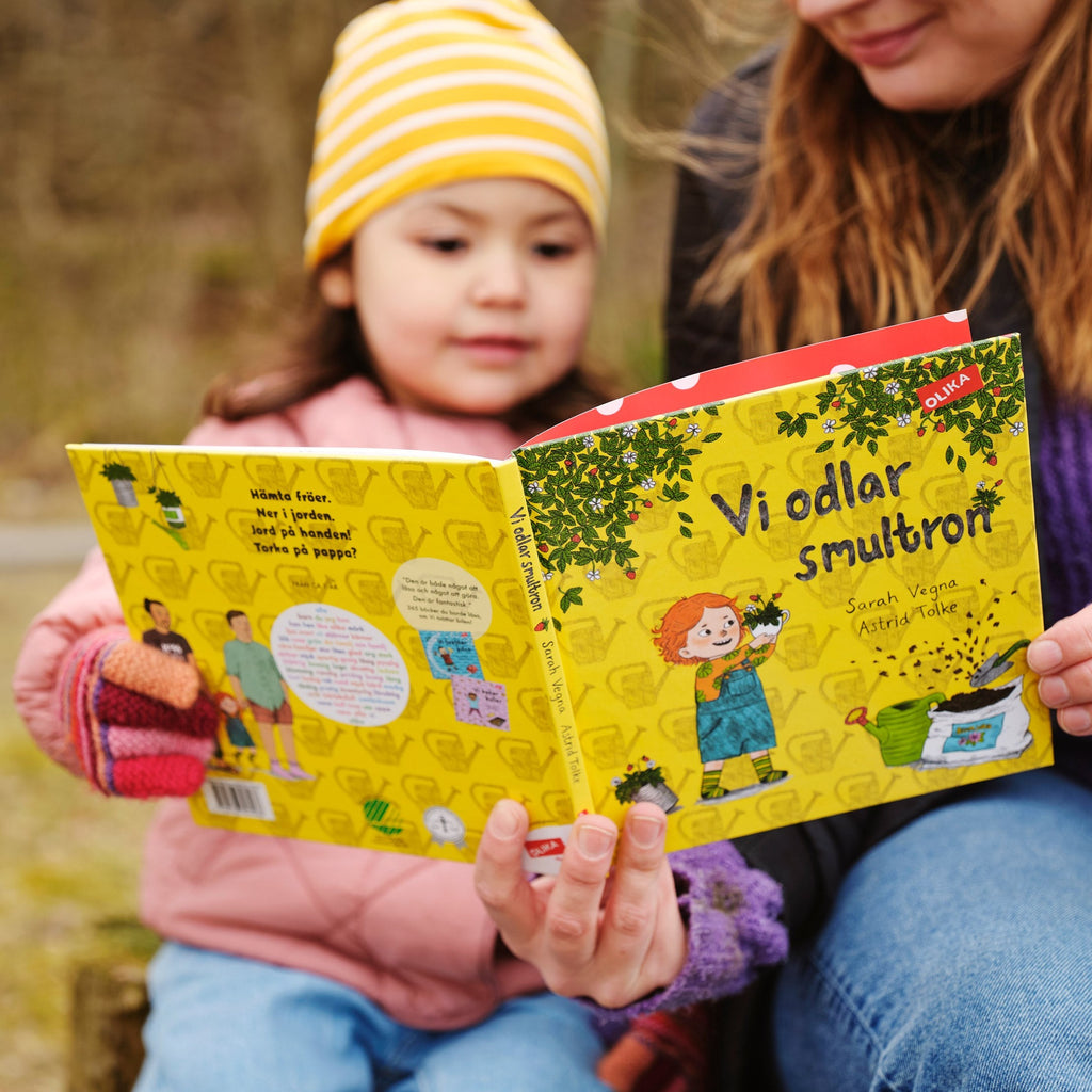 Ett barn och en vuxen sitter utomhus och läser tillsammans i Vi odlar smultron - Bilderbok 1-3 år - OLIKA förlag - Författare: Sarah Vegna - Illustratör: Astrid Tolke