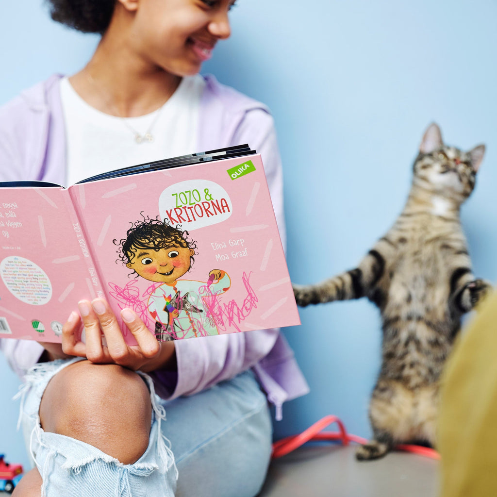 En ungdom ler mot en katt och håller upp en kopia av Zozo och kritorna - Bilderbok 1-3 år - OLIKA förlag - Författare: Elina Garp - Illustratör: Moa Graaf