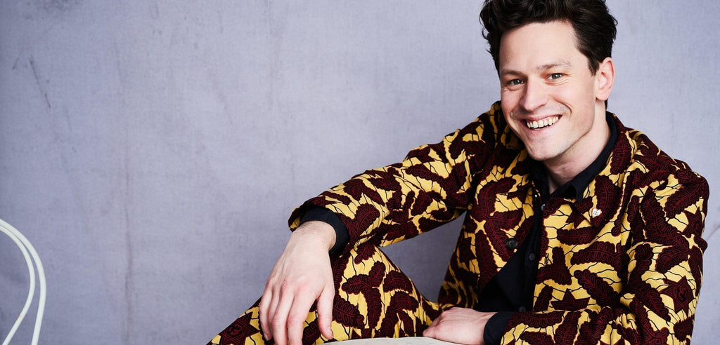  Färgfoto av Henrik Wahlström mot grå vägg. Han sitter på golvet och ler mot kameran. I hörnet syns delvis en stolsrygg