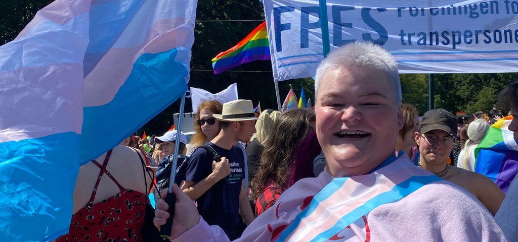 Färgfoto av Louise Sundqvist som deltar i en prideparad, ser in i kameran och håller i en stor transflagga