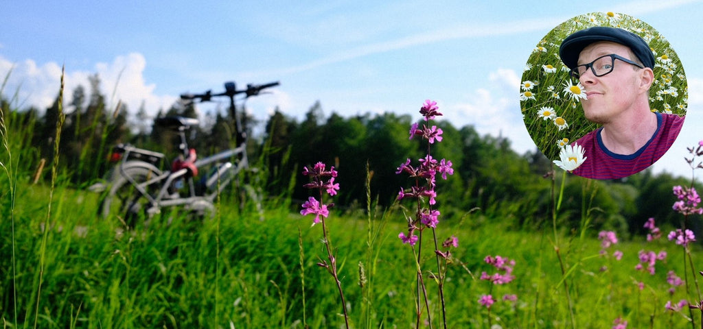 Färgfoto av en cykel som står i gräs under blå sommarhimmel. I övre högra hörn syns ett mindre foto av OLIKAs Leo