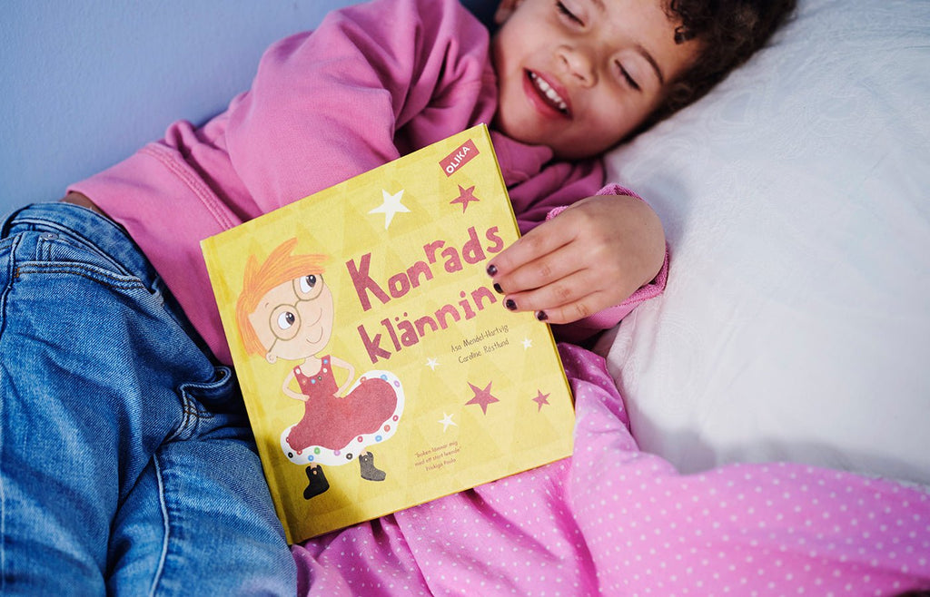 Ett leende barn i jeans, rosa tröja och svart nagellack ligger på en säng & visar bilderboken Konrads klänning för kameran