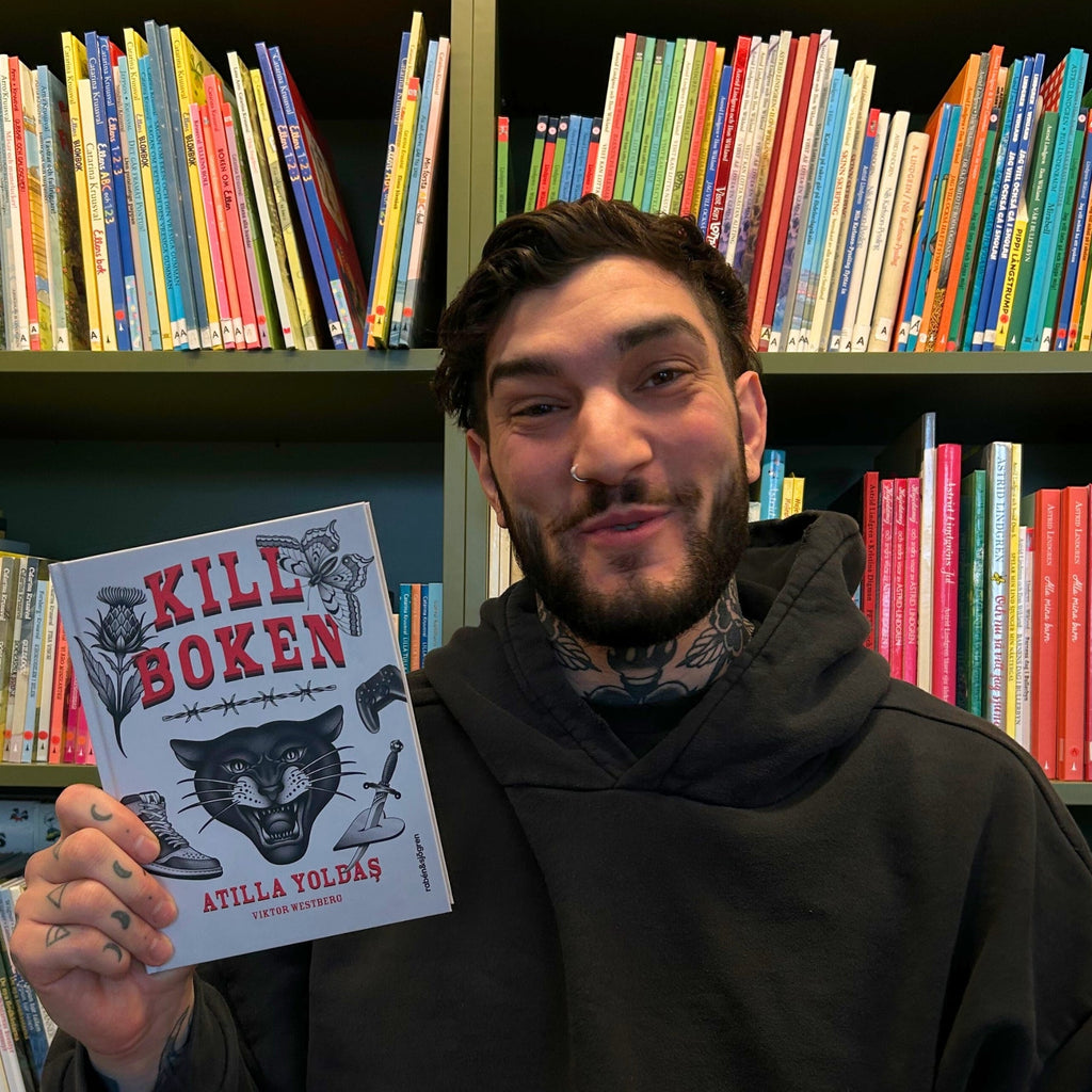 Författaren Atilla Yoldas håller upp sin bok Killboken, i bakgrunden är en bokhylla full med böcker 