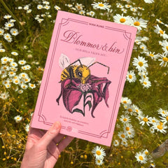 Någon håller i metodboken Blommor och bin och hela vägen dit, på marken syns gräs och blommor