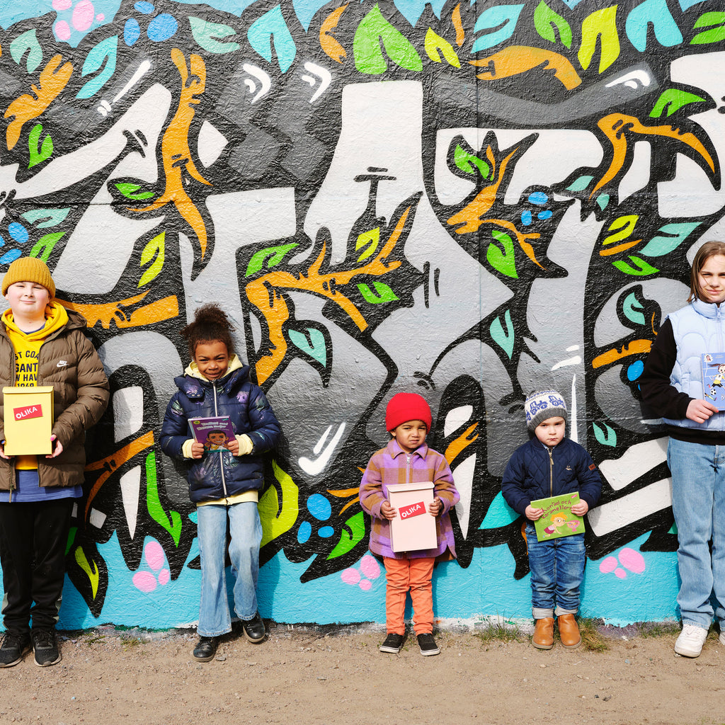 Fem barn i olika åldrar håller i varsin bok och står emot en vägg med graffitti. 