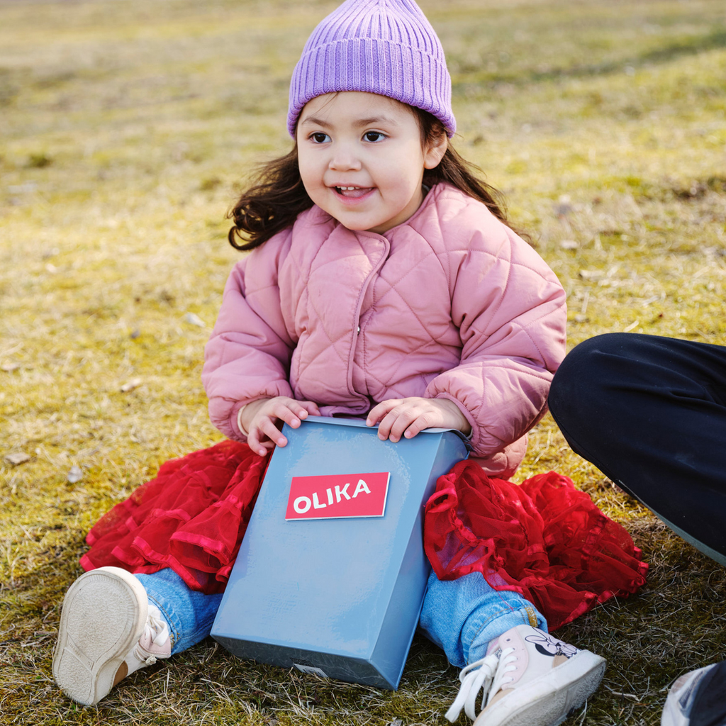 Ett glatt barn sitter på en gräsmatta med en brevlåda i knät, som det står OLIKA på. 