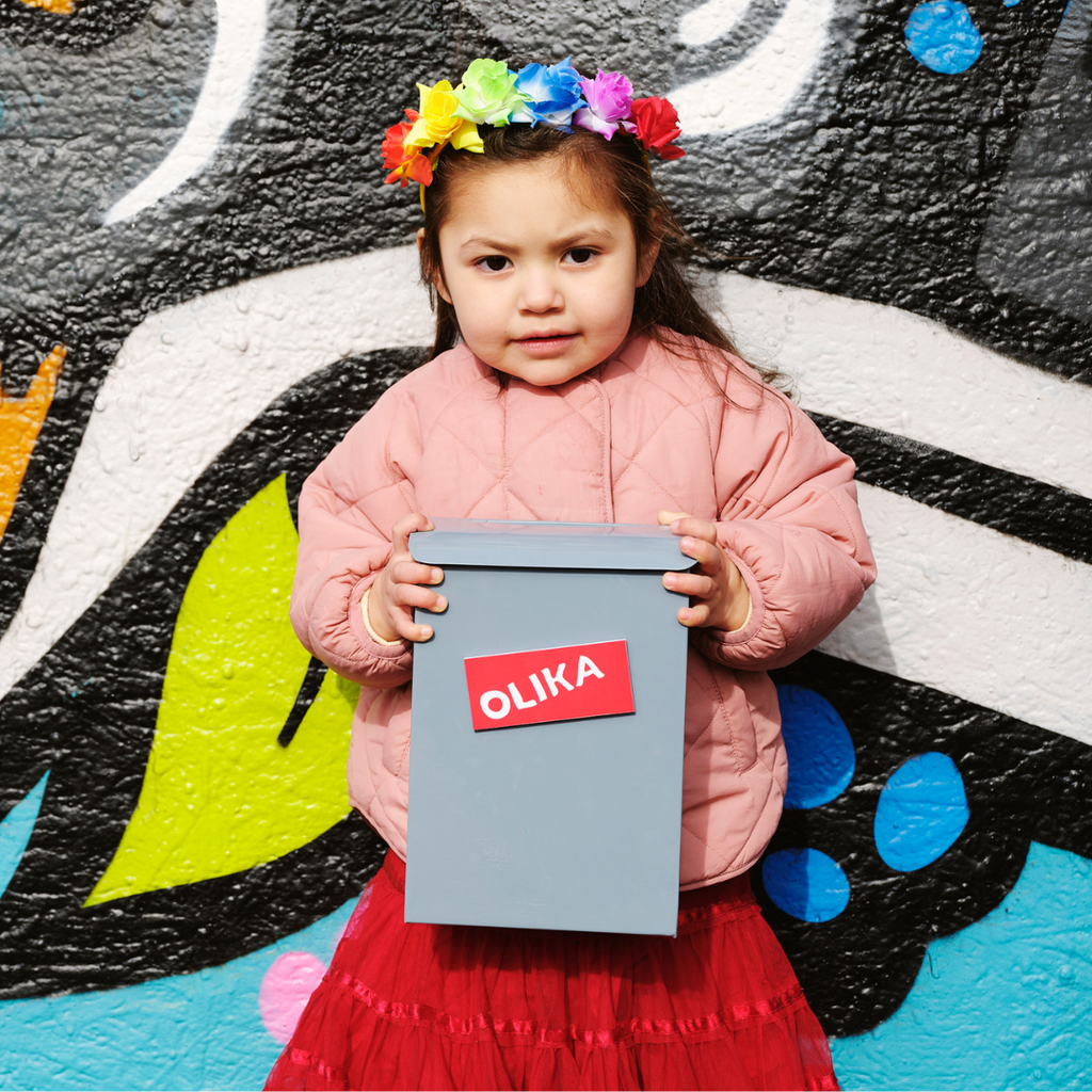 Ett barn med färgglad krans på huvudet står mot en vägg med en brevlåda i händerna. Det står OLIKA på brevlådan. 