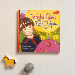 Bilderboken Fest i Sápmi - Feasta Sámis på vit träyta. Under boken ligger en gul leksakshäst i trä och en leksakselefant