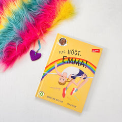 Kapitelboken Flyg högt, Emma! syns på vit trä yta, i hörnet syns en lurvig regnbågsväska med en berlock i form av ett hjärta