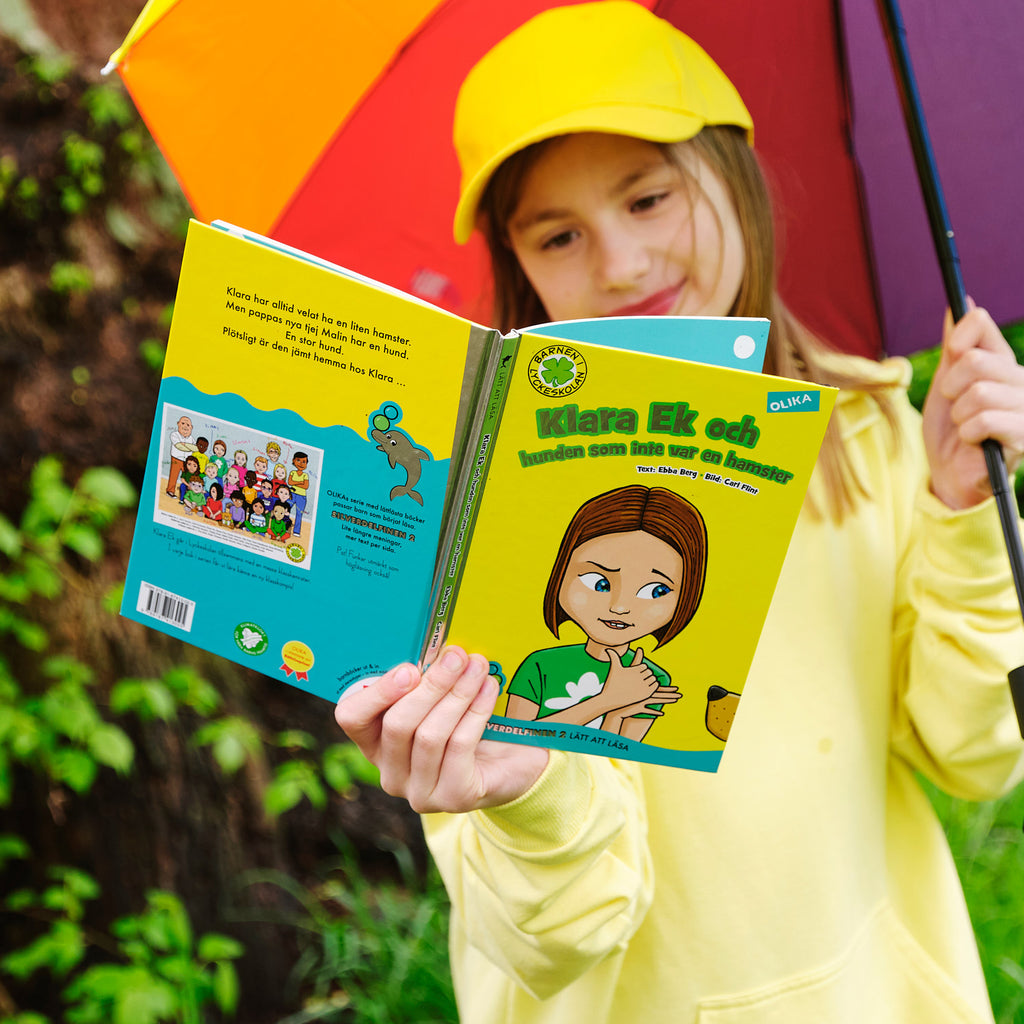Ett barn som håller upp ett regnbågsparaply läser Klara Ek och hunden som inte var en hamster.