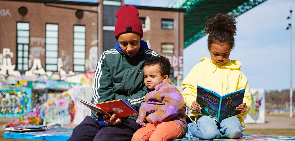 En vuxen och två barn sitter och läser ute med en graffittivägg i bakgrunden. 