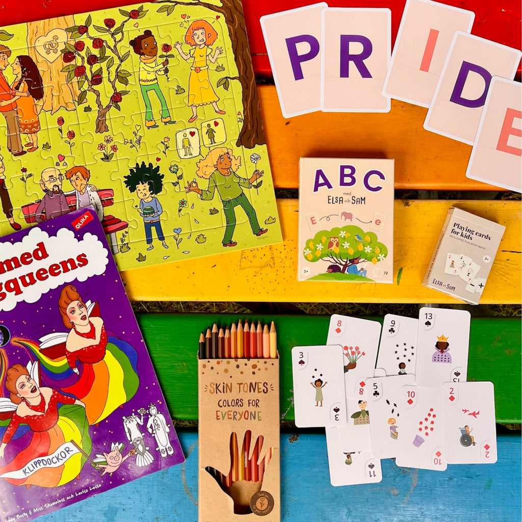 Pussel, spelkort, pennor och ritbok som ingår i paketet syns mot ett regnbågsfärgat bord. 