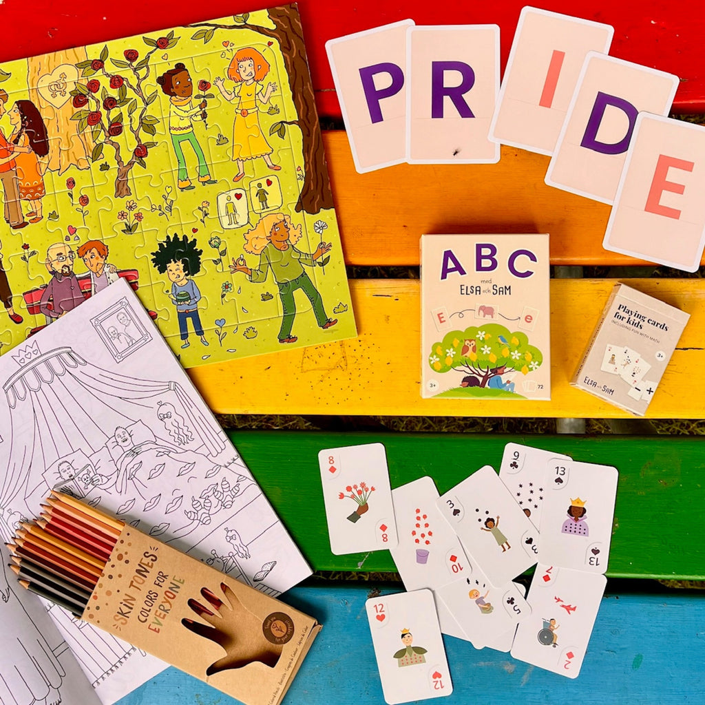 Pussel, spelkort, pennor och ritbok som ingår i paketet syns mot ett regnbågsfärgat bord.
