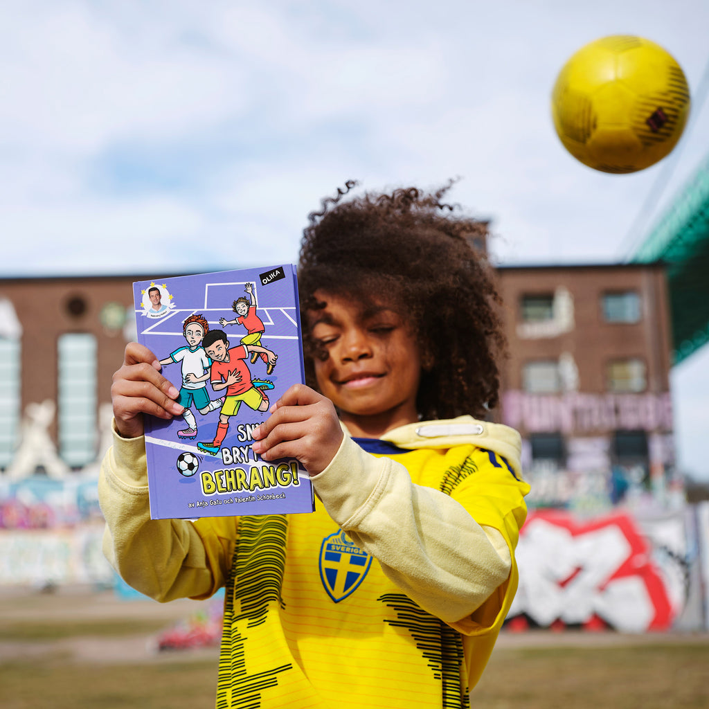 Ett barn som har på sig Sveriges landslag i fotbolls tröja håller upp boken Snygg brytning, Behrang! Uppe i högra hörnet syns en boll flyga i luften. 