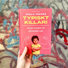 Någon håller upp medtodboken Typiskt killar! mot en vägg full av färgglad graffitti 