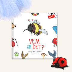 Pekboken Vem är det? på vit yta. I övre hörnet syns ljusblått tyll, i nedre hörnet en virkad nyckelpiga