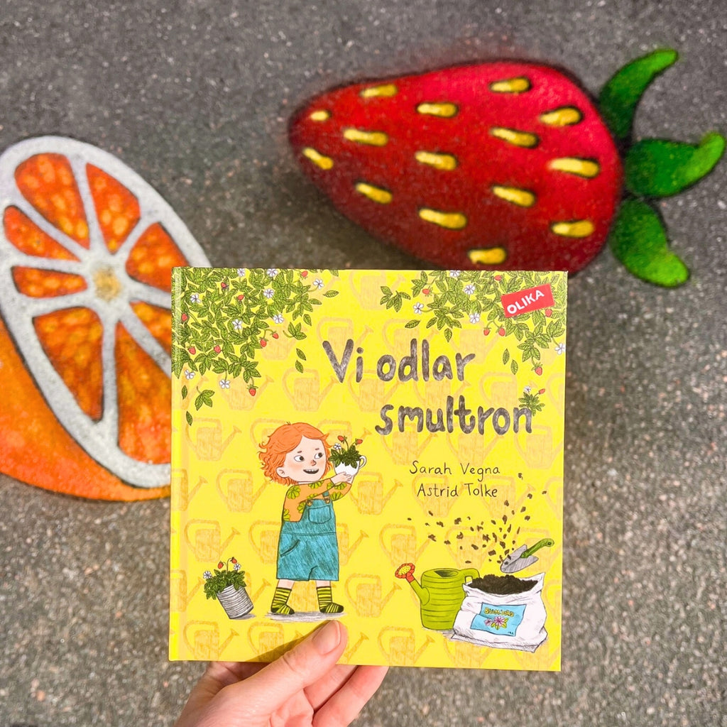 Barnboken Vi odlar smultron hålls upp mot asfalterad bakgrund med gatumålning