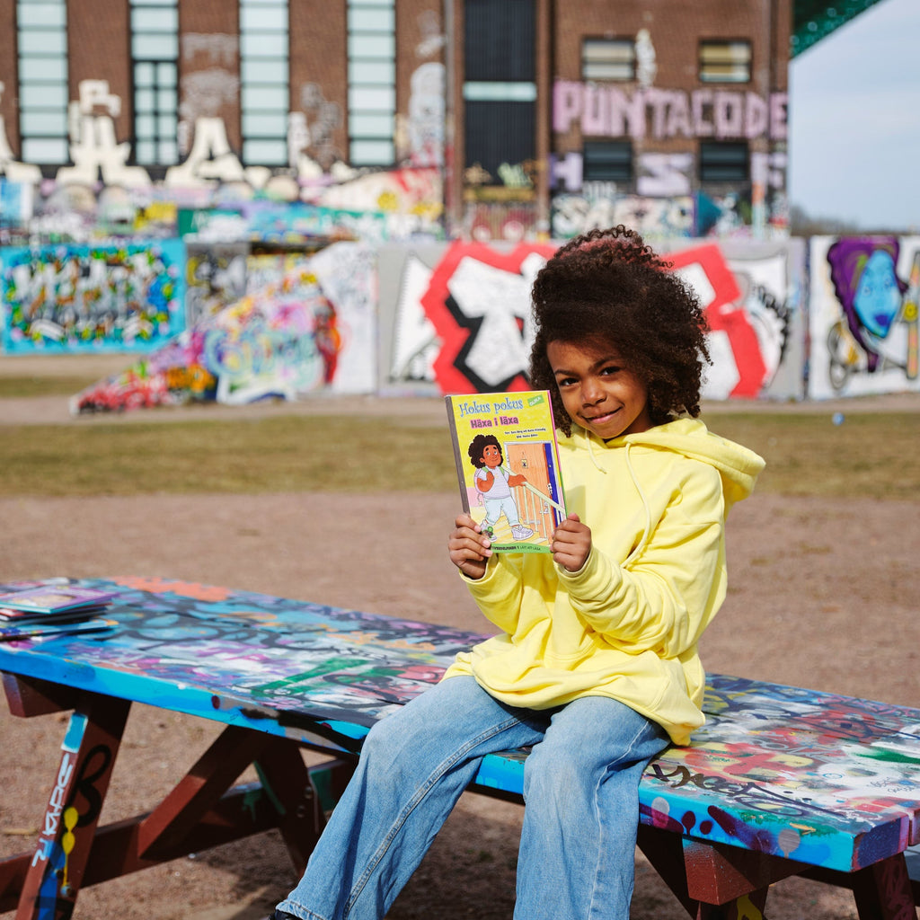 Ett barn sitter på ett picknickbord och håller upp en kopia av Hokus pokus: Häxa i läxa - Lättläst 6-9 år - OLIKA förlag - Författare: Karin Frimodig och Sara Berg - Illustratör: Hanna Böhm