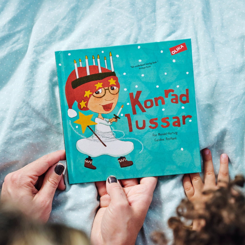 En person med svarta naglar visar ett barn framsidan av Konrad lussar - Bilderbok 1-3 år - OLIKA förlag - Författare: Åsa Mendel-Hartvig - Illustratör: Caroline Röstlund