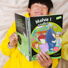 Ett barn ligger bredvid någon i gul regnjacka på en säng och läser lätt att läsa boken Malva i stallet