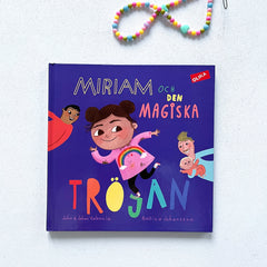 Bilderboken Miriam och den magiska tröjan på vit träyta. Ovanför boken ligger ett färgglatt halsband