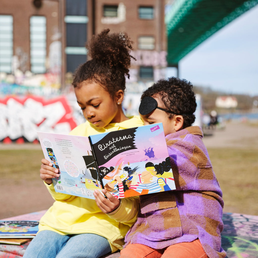 Två barn i färgglada kläder sitter utomhus och läser i Piraterna & förtrollningen - Bilderbok 3-6 år - OLIKA förlag - Författare: Karin Frimodig och Sara Berg - Illustratör: Maria Poll