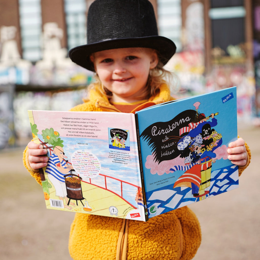 Ett leende barn i gul tröja håller upp en kopia av Piraterna & vinterjakten - Bilderbok 3-6 år - OLIKA förlag - Författare: Karin Frimodig och Sara Berg - Illustratör: Maria Poll