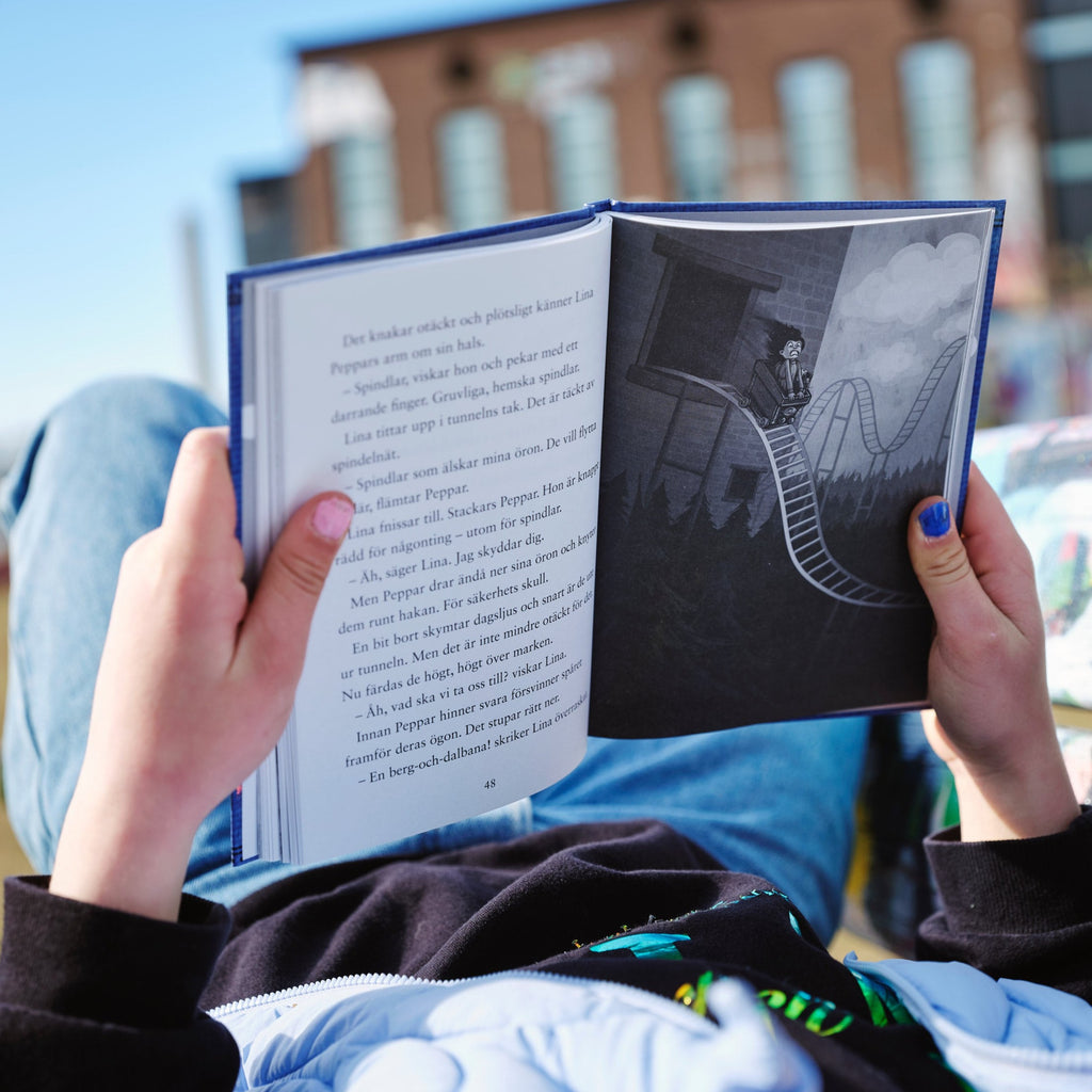 Ett barn ligger utomhus och läser boken Uppdrag: Skrämmarna - Kapitelbok 6-9 år - OLIKA förlag - Författare: Cecilia Rihs - Illustratör: Silvy Strand