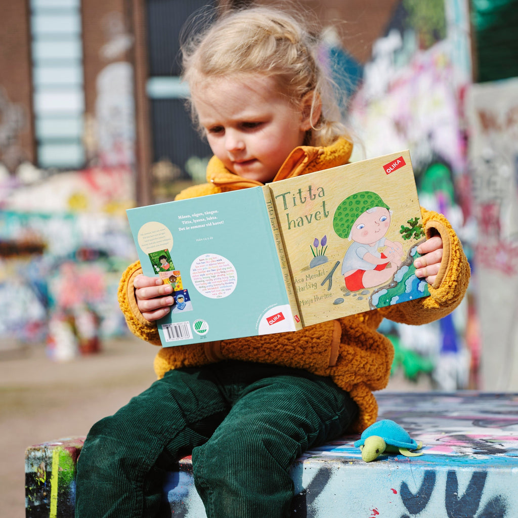 Ett barn i gröna byxor och gul tröja bläddrar i Titta havet! - Bilderbok 0-2 år - OLIKA förlag - Författare: Åsa Mendel-Hartvig - Illustratör: Maija Hurme