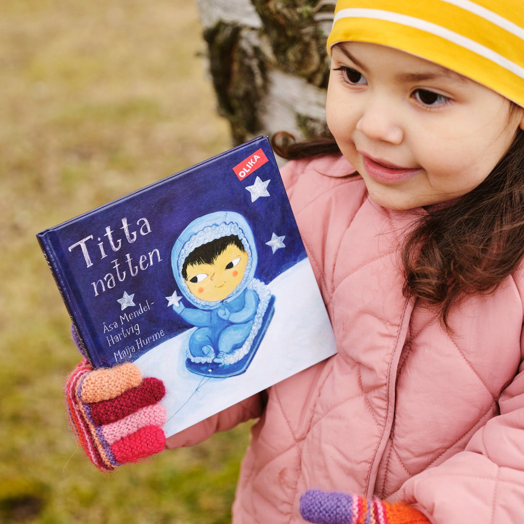 Ett barn i rosa jacka och gulrandig mössa håller upp en kopia av Titta natten! - Bilderbok 0-2 år - OLIKA förlag - Författare: Åsa Mendel-Hartvig - Illustratör: Maija Hurme