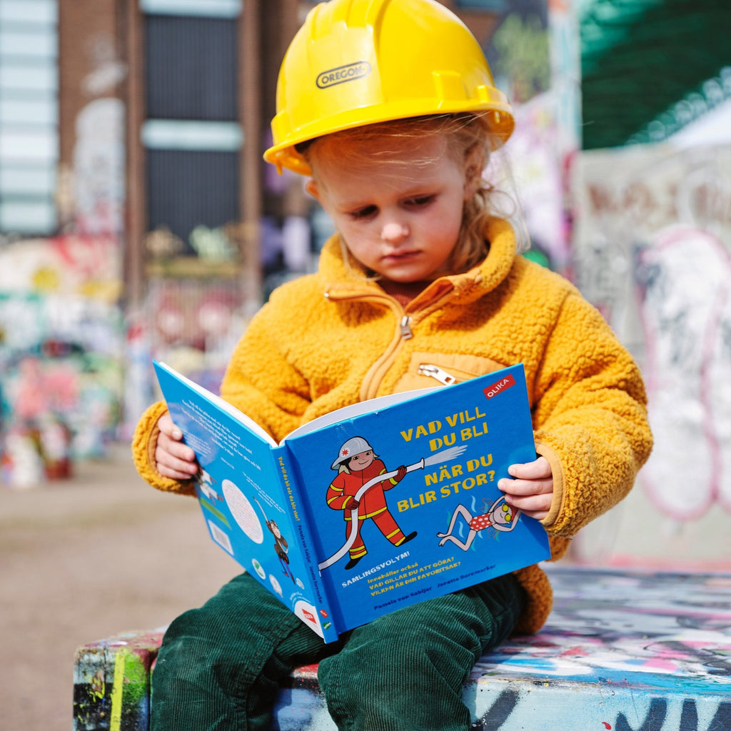 Ett barn i gul bygghjälm som bläddrar i Vad vill du bli när du blir stor? - 3-6 år - OLIKA förlag - Författare: Pamela von Sabljar - Illustratör: Janette Bornmarker