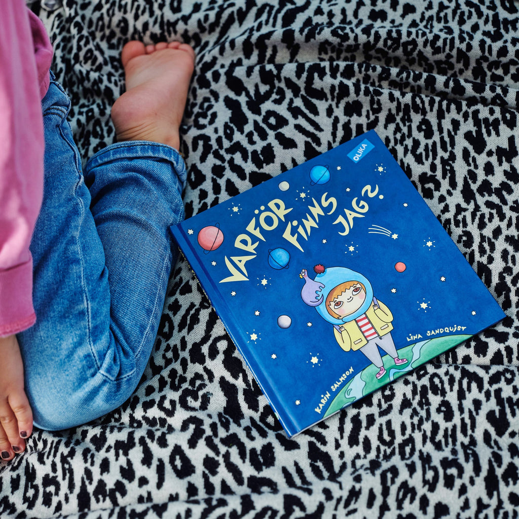 Ett barn sitter ovanpå en säng bredvid en kopia av Varför finns jag? - Bilderbok 1-3 år - OLIKA förlag - Författare: Karin Salmson - Illustratör: Lina Sandquist