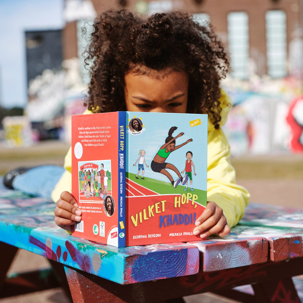 Ett barn ligger ovanpå ett målat picknickbord och läser i Vilket hopp, Khaddi! - Baserad på friidrottaren Khaddi Sagnias barndom - Kapitelbok 6-9 år - OLIKA förlag - Författare: Behrang Behdjou - Illustratör: Micaela Favilla